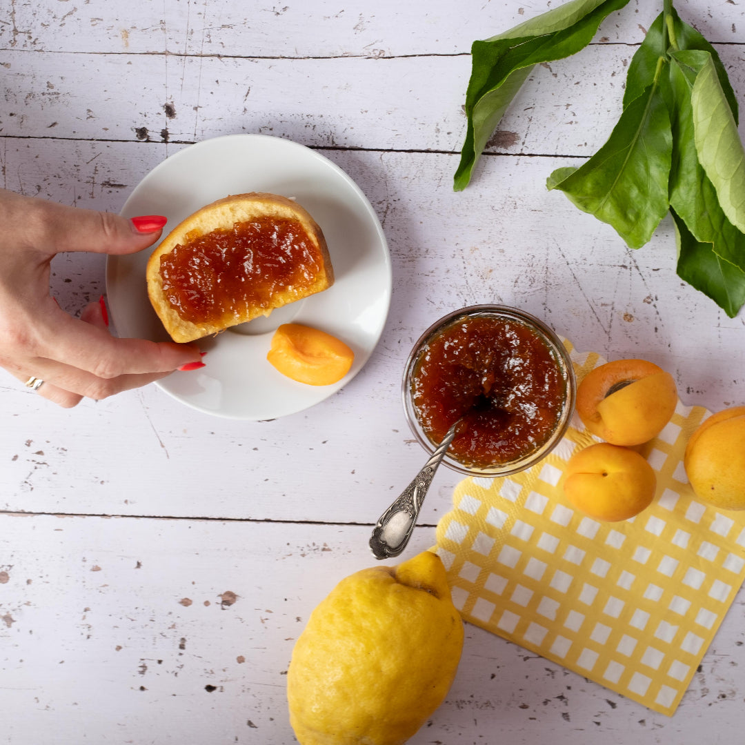 Aprikosen-Fruchtaufstrich von Kuchentratsch auf Tisch mit frischen Aprikosen und einer Zitrone - Eine Hand greift nach einem Brioche mit Marmelade