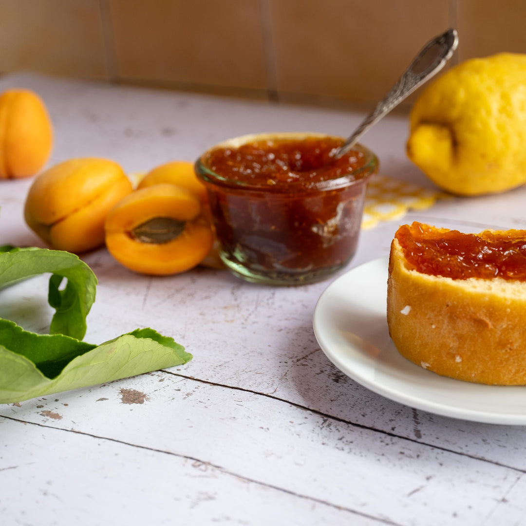 Aprikosen-Fruchtaufstrich von Kuchentratsch auf Tisch mit frischen Aprikosen und einer Zitrone, daneben ein Teller mit einem mit Marmelade bestrichenem Brioche