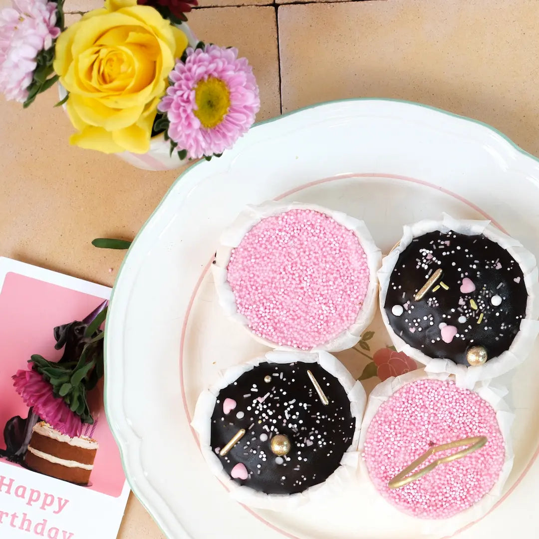 Vier Geburtstagsmuffins von Kuchentratsch auf einem Teller: zwei mit rosa Streuseln, zwei mit Schokoglasur. In einem Muffin steckt eine goldene Herz-Wunderkerze.