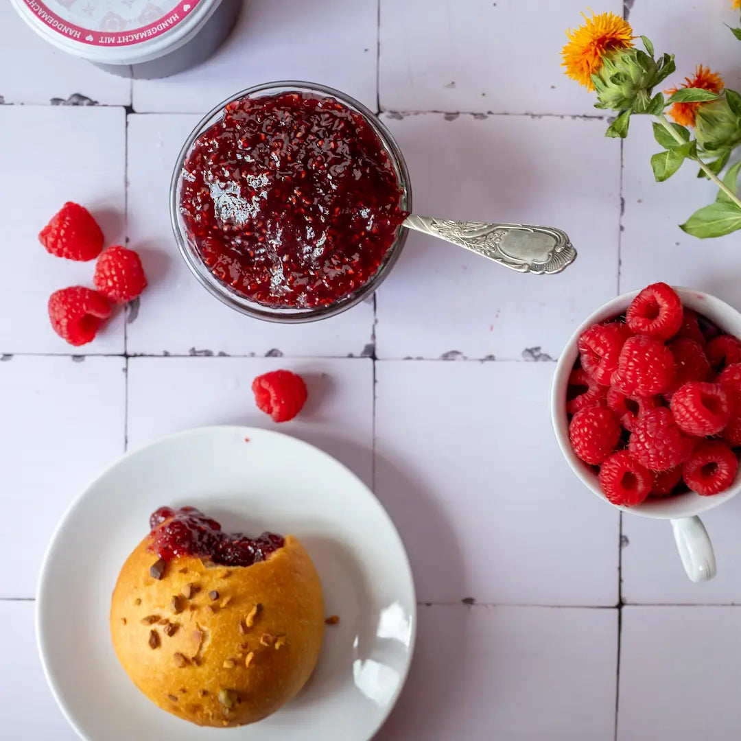 Himbeer-Fruchtaufstrich mit frischen Himbeeren in einer Schale. Im Hintergrund ein Brioche bestrichen mit dem Fruchtaufstrich.