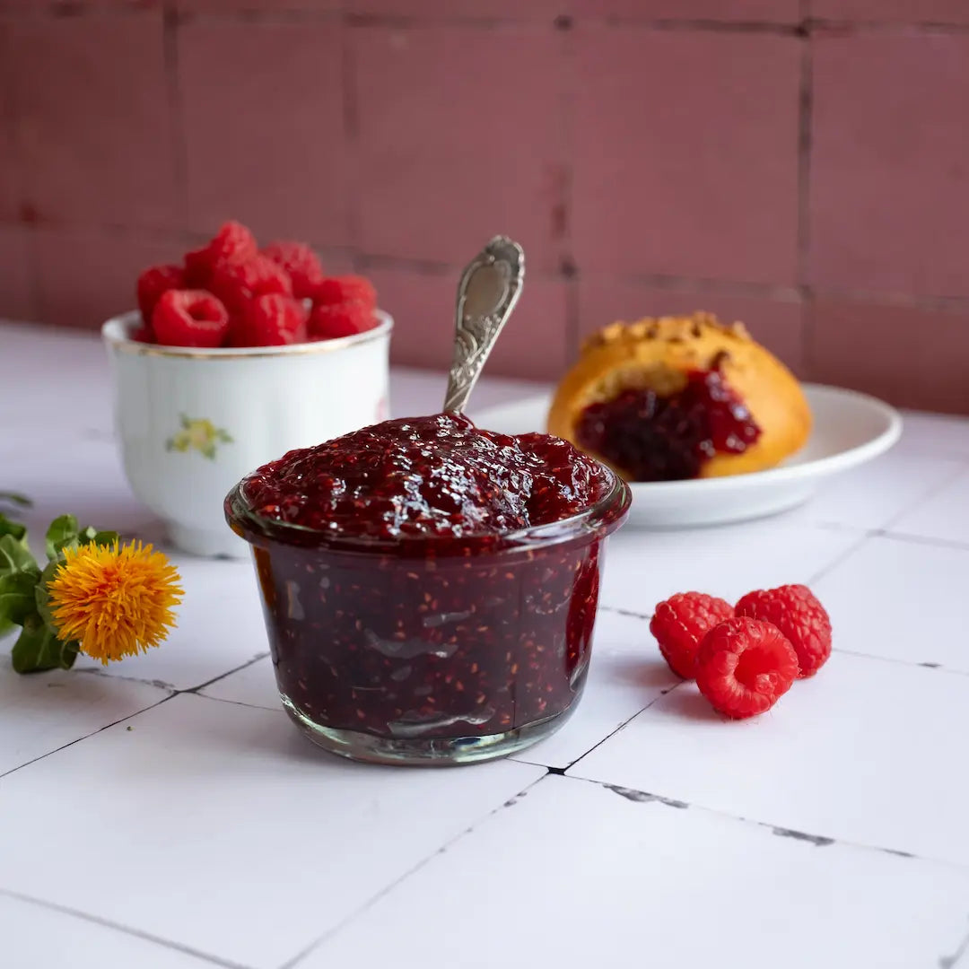 Himbeer-Fruchtaufstrich mit frischen Himbeeren in einer Schale. Im Hintergrund ein Brioche bestrichen mit dem Fruchtaufstrich.