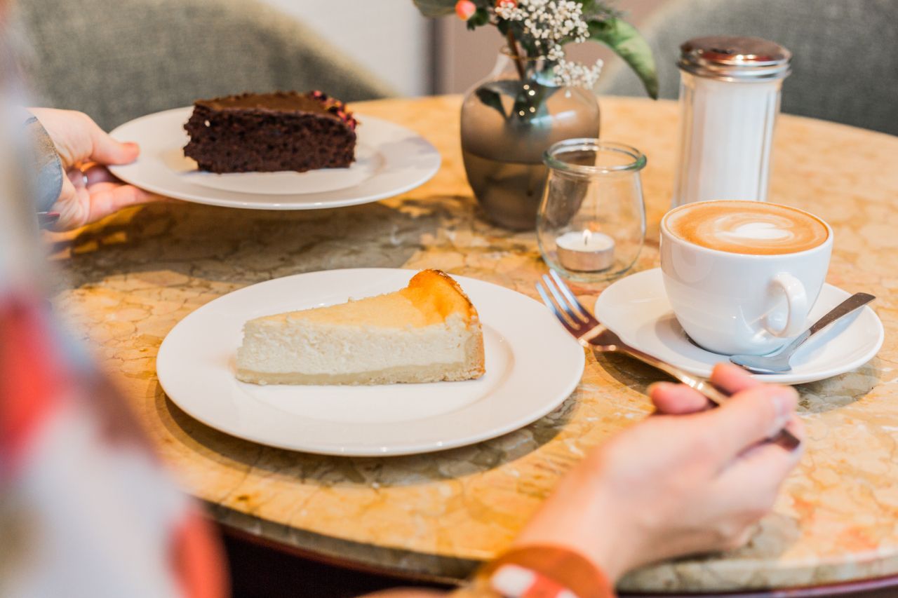 Bild aus dem Kuchentratsch Cafe, eine Person sitzt an einem Tisch und isst ein Stück Käsekuchen und trinkt einen Cappuccino