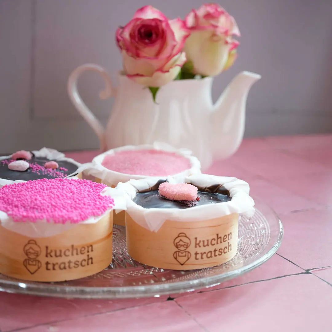 Die Kuchentratsch Amoretörtchen im 4er Set dekoriert mit handegmachter Herzchenverzierung fotografiert auf einem rosa Tisch mit rosen im Hintergrund