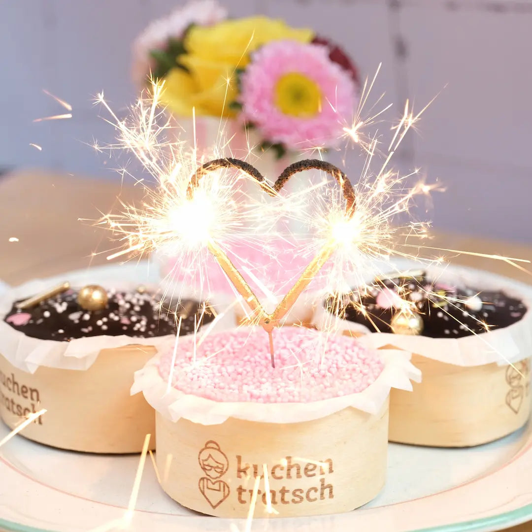 Brennende Herz-Wunderkerze auf einem Muffin mit rosa Streuseln. Dahinter stehen weitere Geburtstagsmuffins.