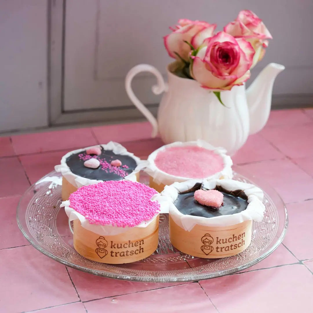 Die Kuchentratsch Valentinstagstörtchen im 4er Set dekoriert mit handegmachter Herzchenverzierung fotografiert auf einem rosa Tisch mit rosen im Hintergrund